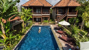 A swimming pool with a person diving, surrounded by lush greenery and two wooden buildings with tiled roofs. - JAVA WOODEN VILLA (Siem Reap)