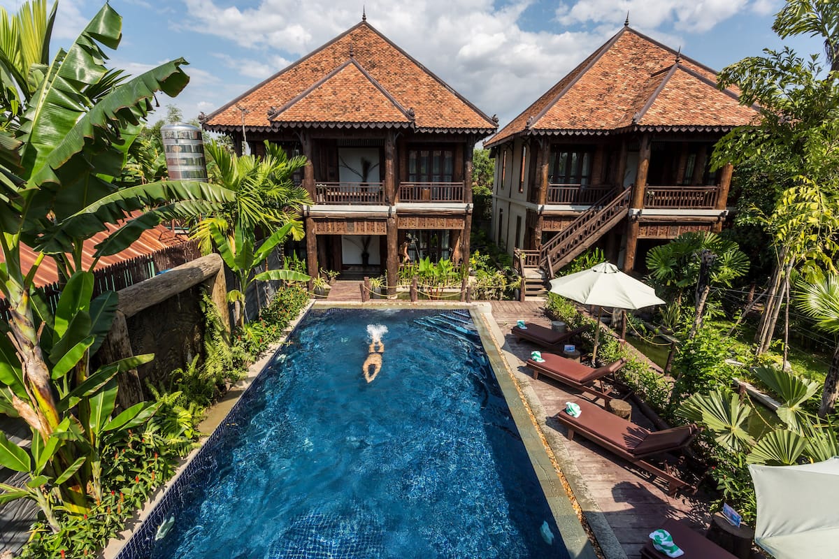 A swimming pool with a person diving, surrounded by lush greenery and two wooden buildings with tiled roofs.