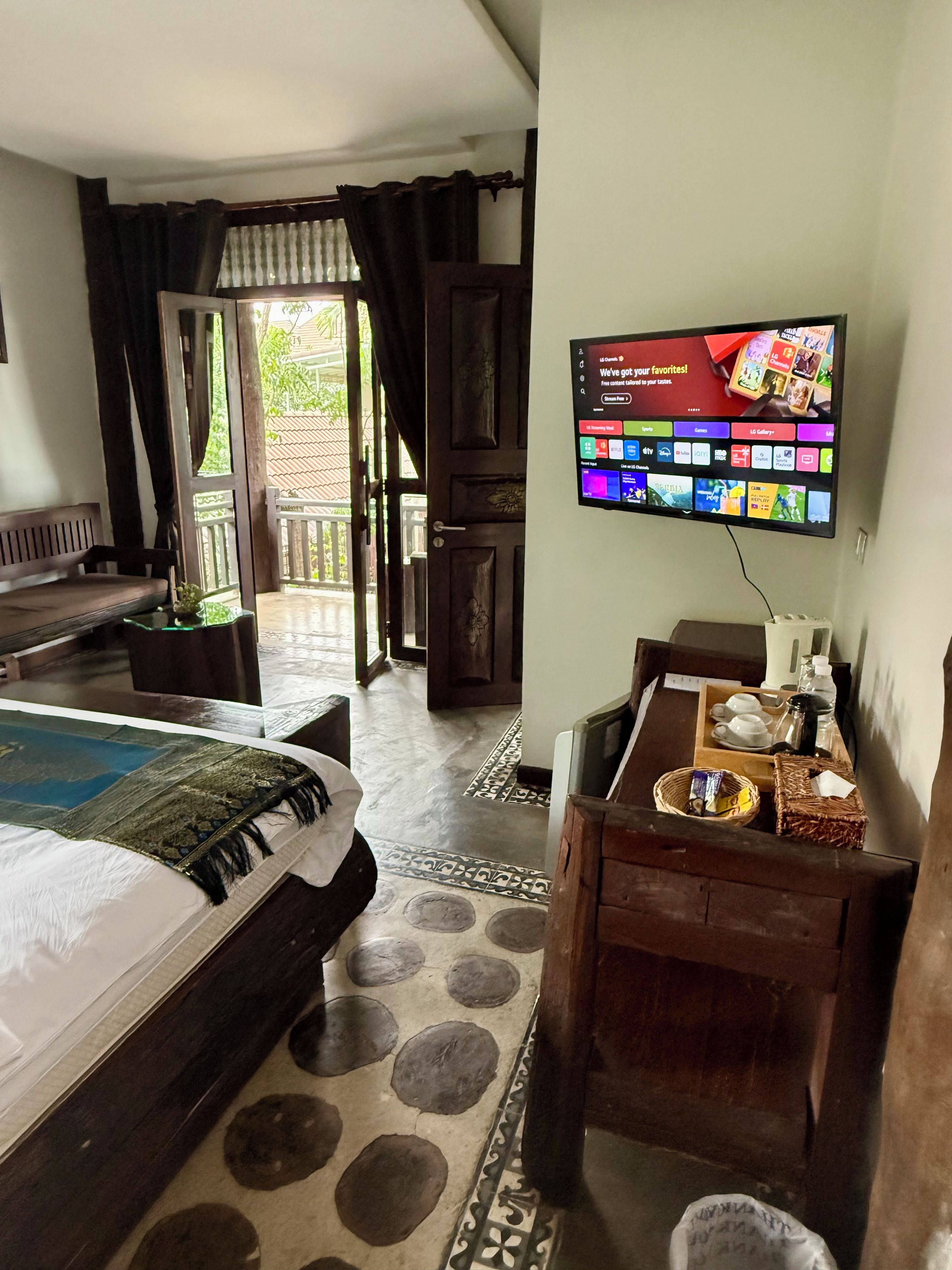 A hotel room with a bed, a TV mounted on the wall, a small table with a tray of breakfast items, and a view of the outside through a door.