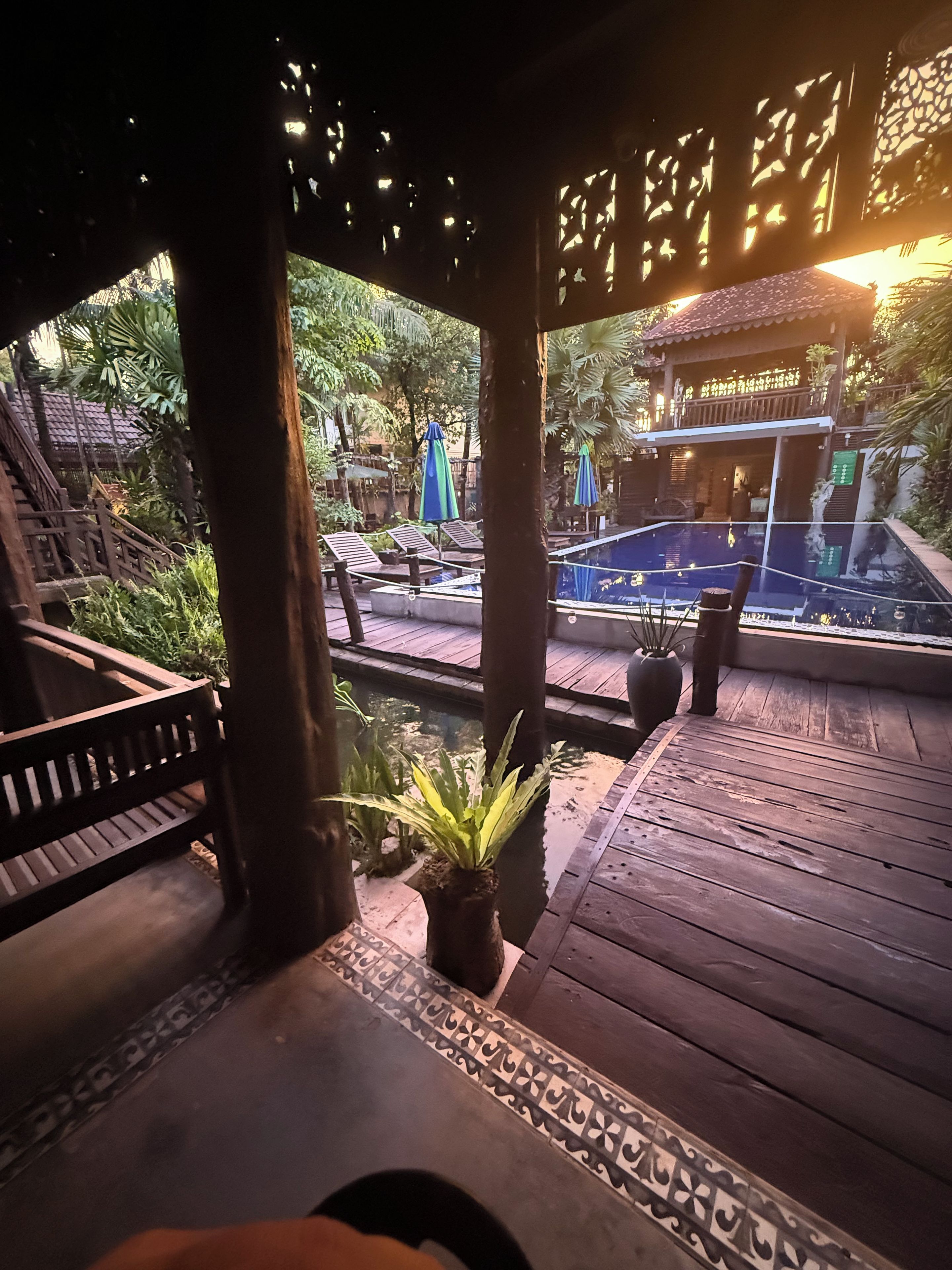A wooden deck with a pool, lounge chairs, and a covered area with a view of a building in the background.