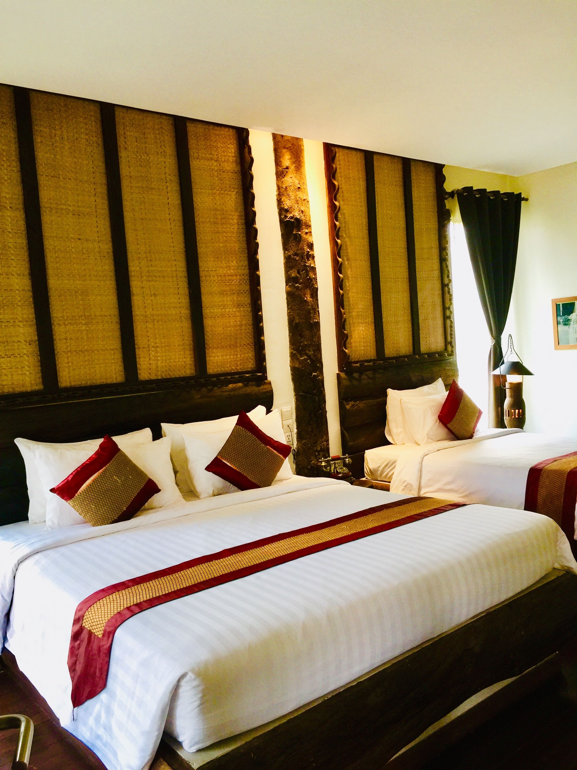 Une chambre d’hôtel avec deux lits, une grande fenêtre avec des rideaux, une tête de lit en bois et une table de chevet avec une lampe.