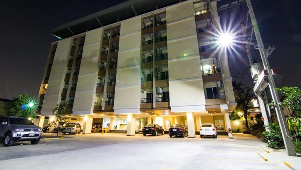 A multi-story hotel with a modern design, featuring a parking area with several cars and a well-lit entrance. - Dae Ma Place (Nonthaburi)