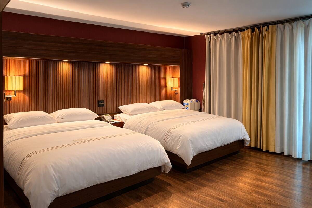A hotel room with two beds, wooden headboard, bedside lamps, and a large window with curtains.
