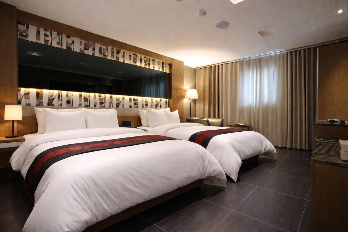 A hotel room with two beds, a large mirror, a flat-screen TV, a desk, and a window with curtains.