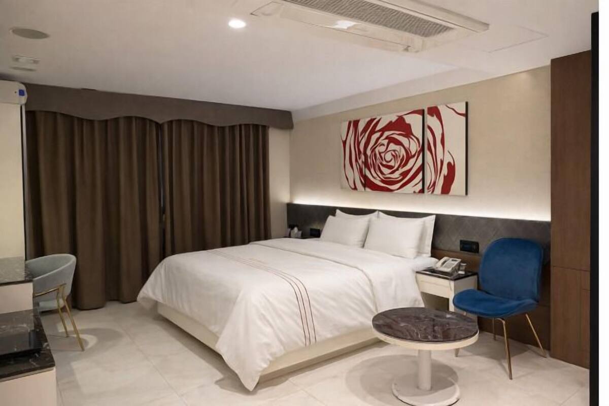 A modern hotel room with a large bed, a desk, a chair, a round table, and a wall-mounted artwork.