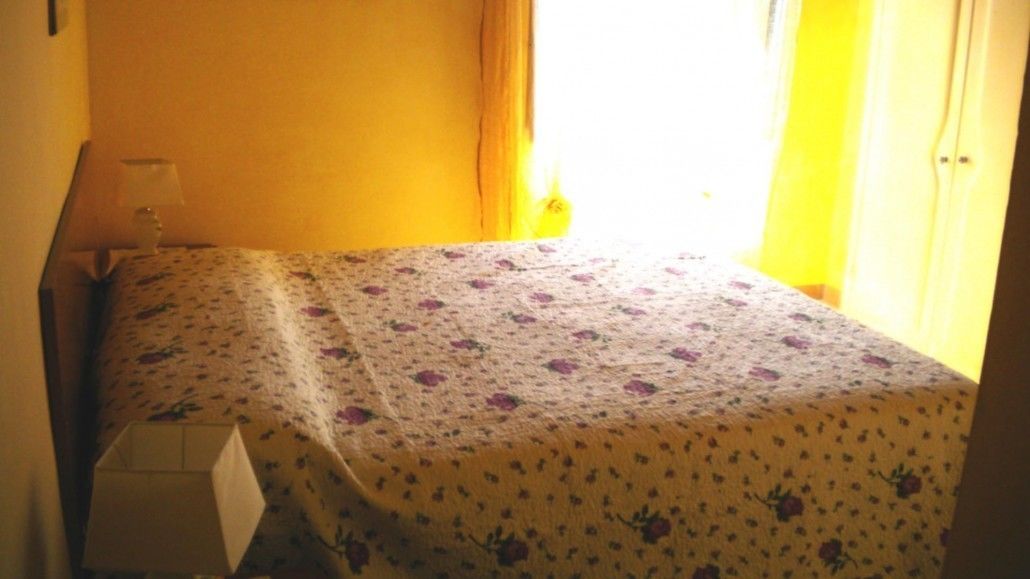 A small, simple room with a bed, a bedside table, and a window with curtains.