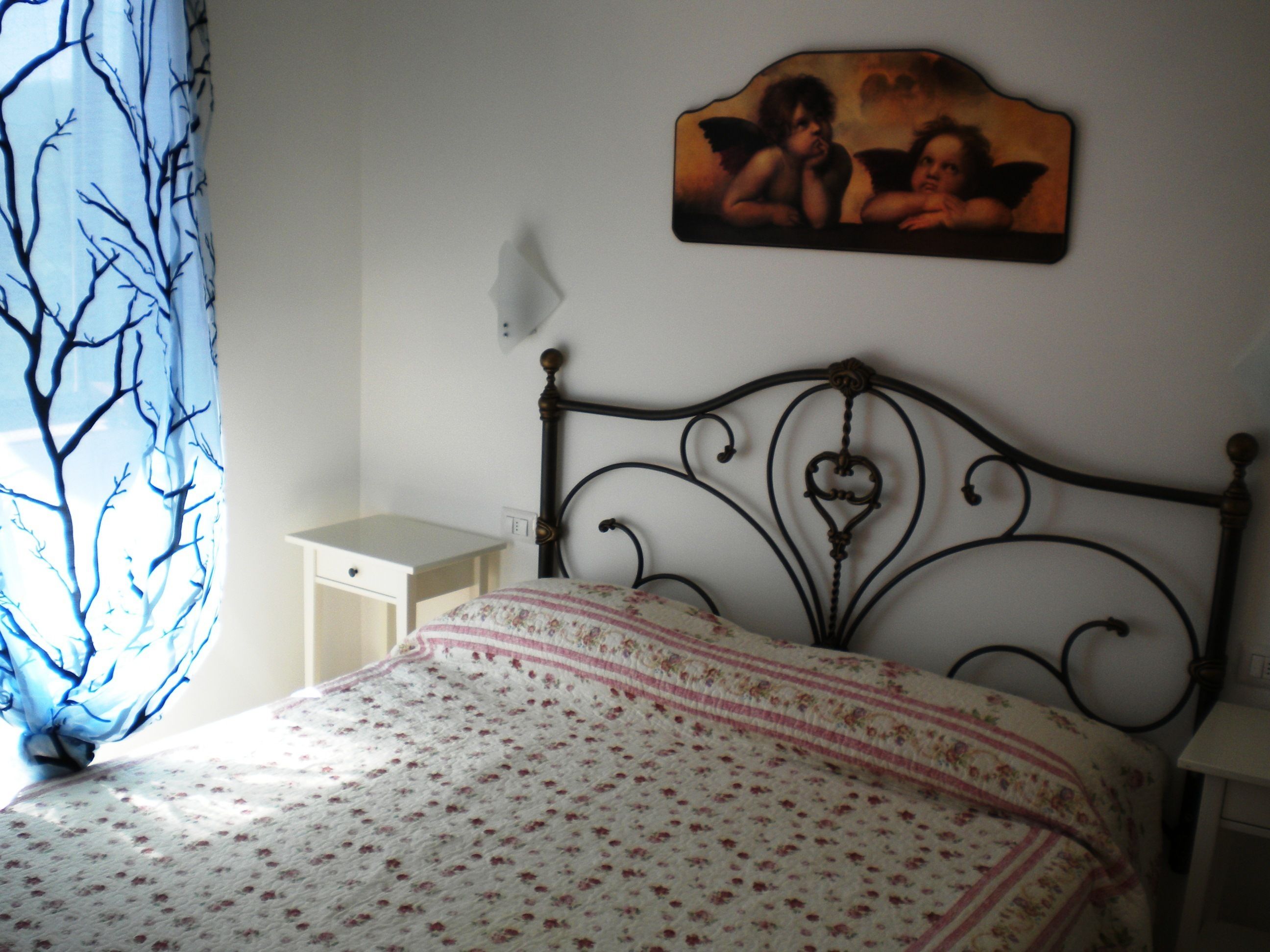 A bedroom with a bed, a nightstand, a wall-mounted picture, and a window with a blue curtain.