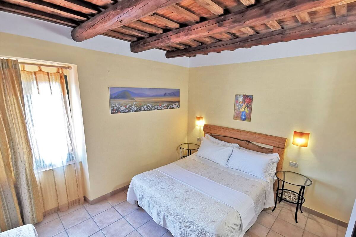 A bedroom with a wooden ceiling, a bed with white linens, two bedside tables, a painting on the wall, and a window with curtains.