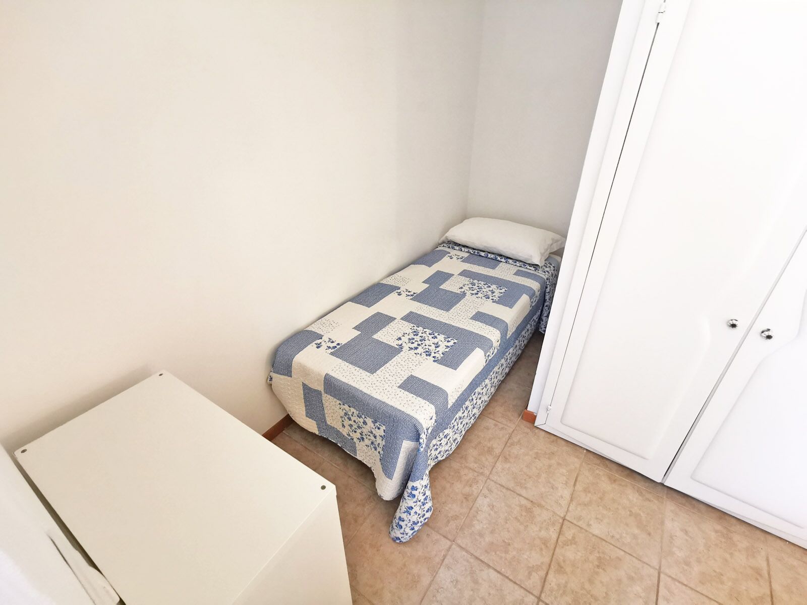 A small, single-bed room with a quilted bedspread, a white cabinet, and a door.