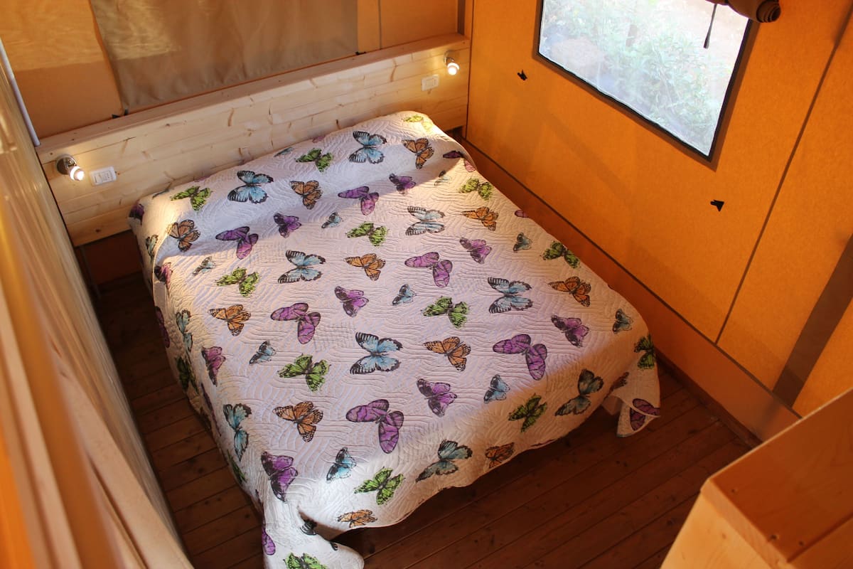 A small, cozy room with a bed covered in a butterfly-patterned quilt, wooden walls, and a window with a view of greenery.