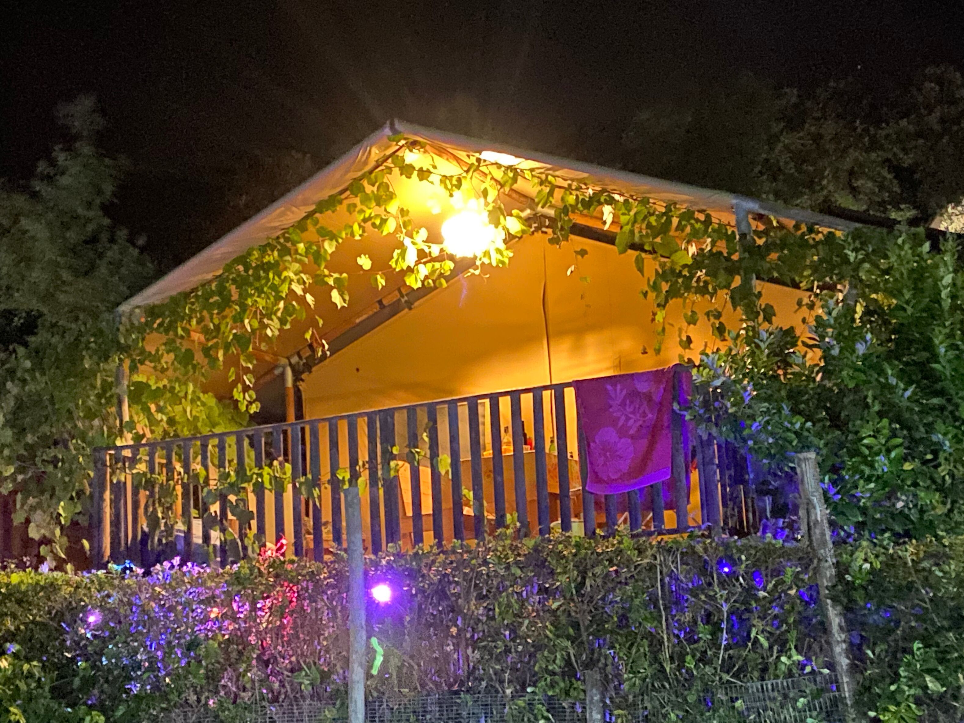 Una estructura parecida a una tienda de campaña con un balcón, rodeada de follaje e iluminada con luz cálida.