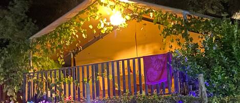 Una estructura parecida a una tienda de campaña con un balcón, rodeada de follaje e iluminada con luz cálida.
