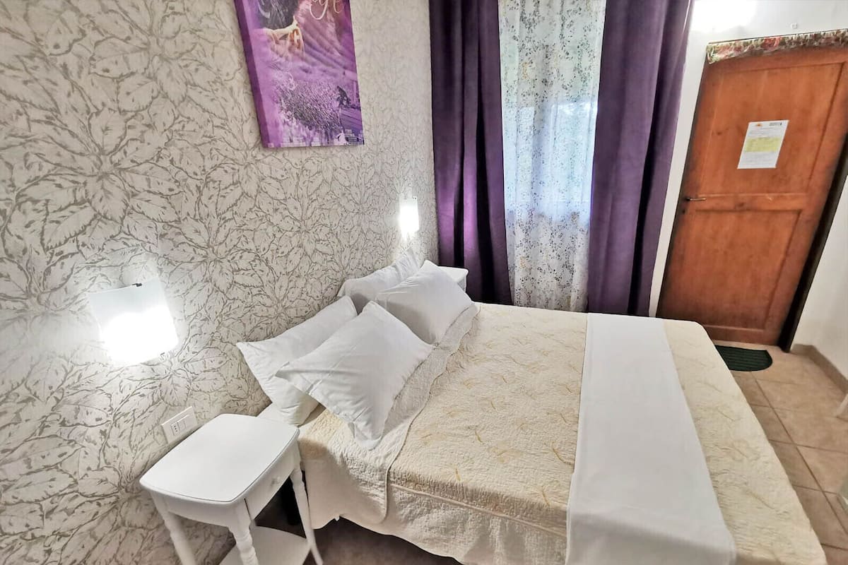 A bedroom with a bed, a small white table, a purple wall decoration, and a wooden door.
