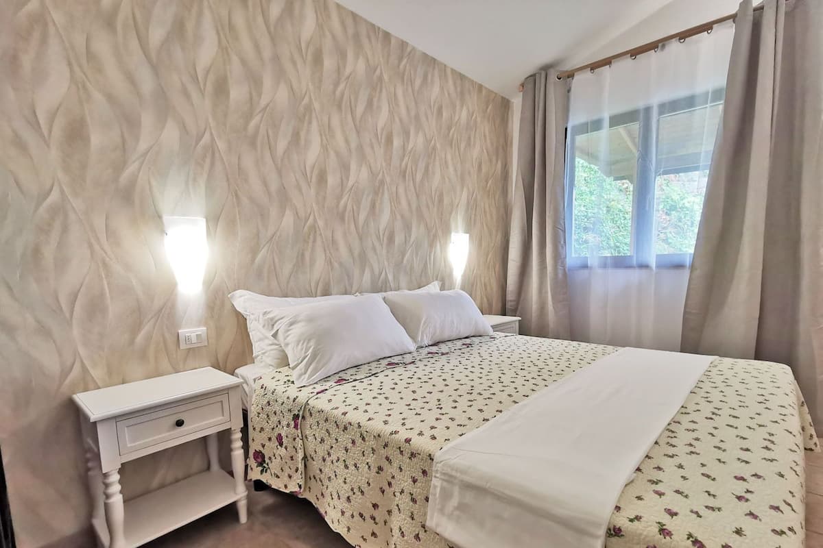 A neatly made bed with a floral bedspread, a bedside table with a lamp, and a window with curtains.