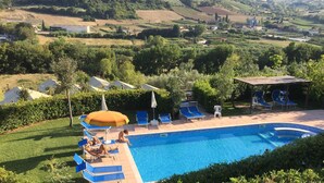 A swimming pool with blue tiles, surrounded by blue lounge chairs and umbrellas, set against a backdrop of green hills and buildings. - Il Girasole (Massignano)