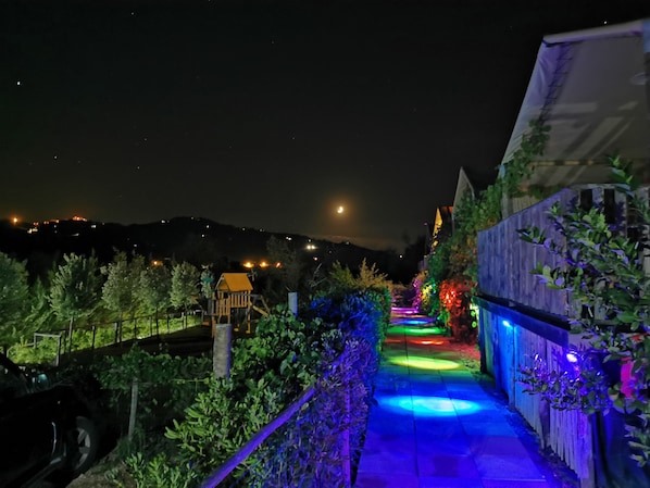 Un sendero en un jardín nocturno con iluminación colorida, una casita de juegos de madera y vistas a colinas distantes.