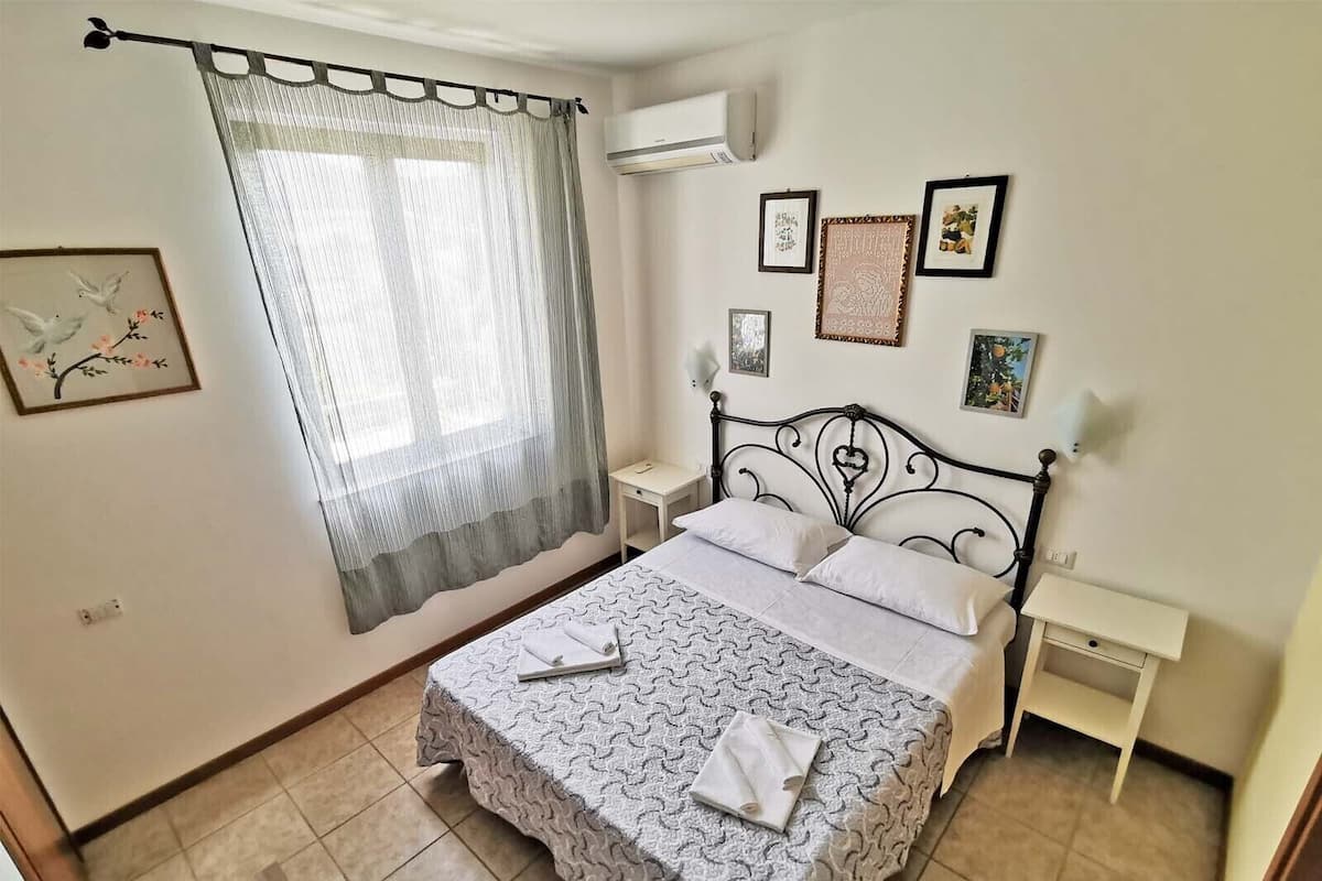 A neatly arranged bedroom with a bed, a bedside table, a nightstand, and a window with sheer curtains.
