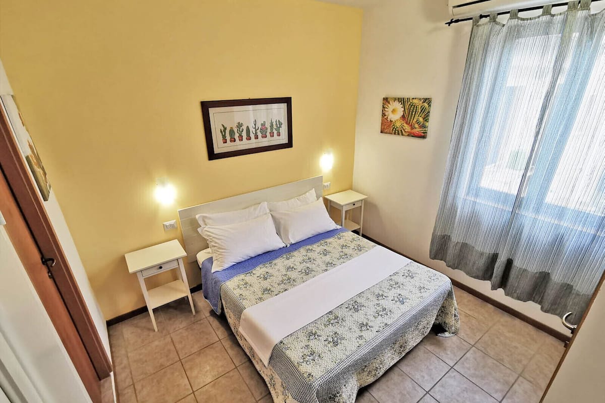 A bedroom with a bed, two bedside tables, a nightstand, a framed cactus artwork, and a window with curtains.