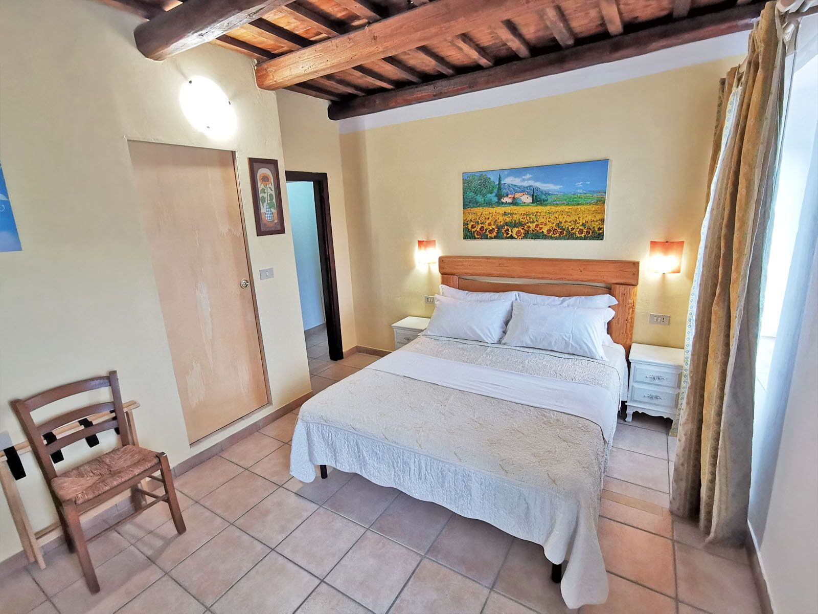 A bedroom with a bed, wooden headboard, two bedside tables, a nightstand, a chair, a painting, and a door.