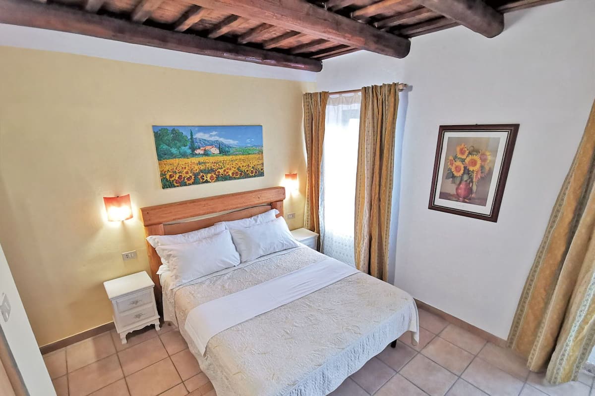 A bedroom with a wooden headboard, a painting of a field, a framed picture of sunflowers, and a nightstand with a lamp.