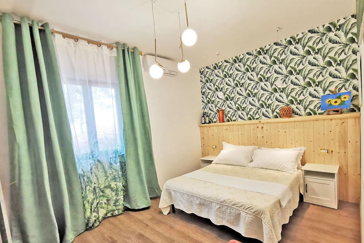A bedroom with a bed, nightstands, a wooden headboard, a wall with a tropical leaf pattern, and a window with green curtains.