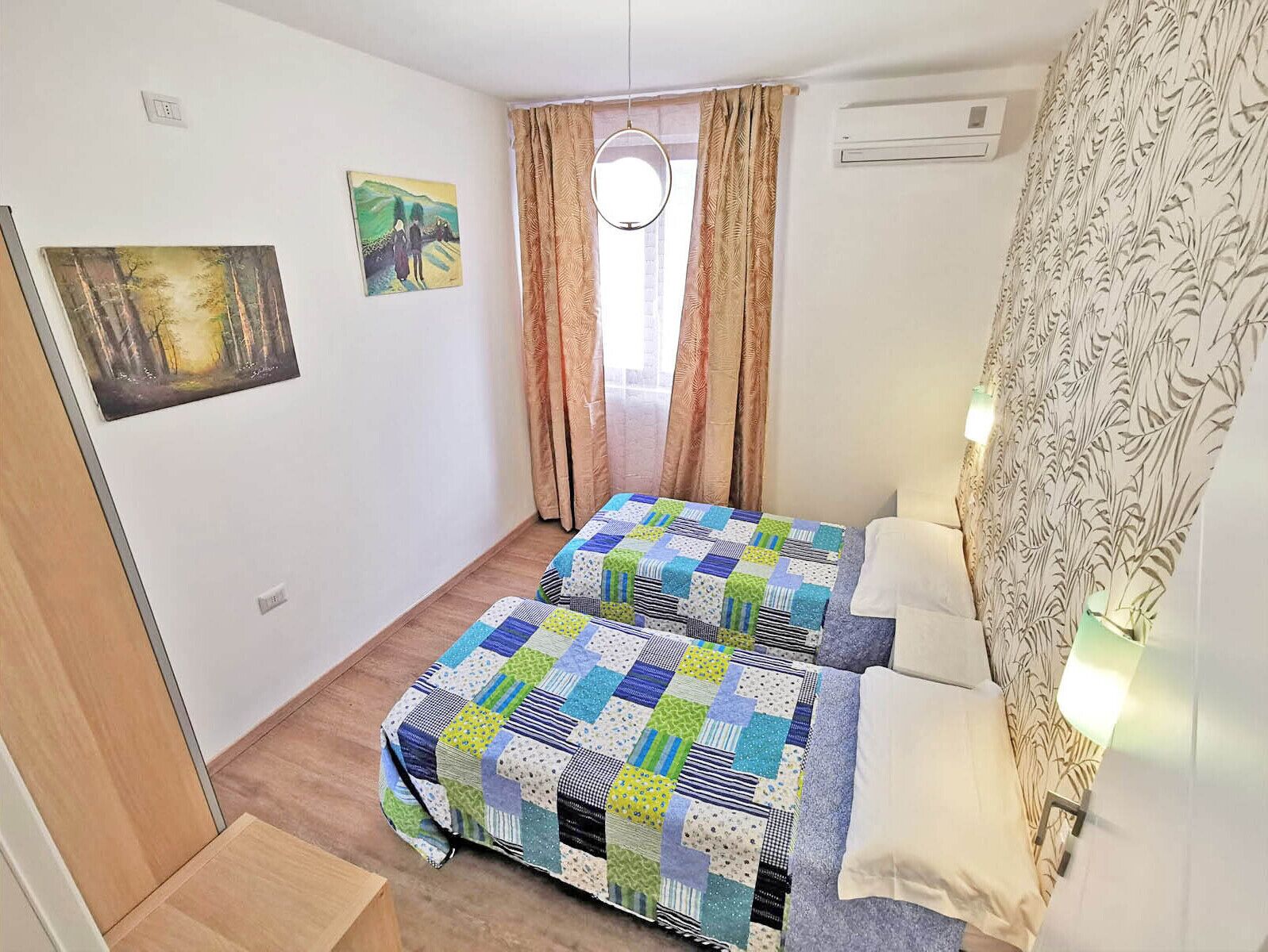 A small, clean room with two beds, a window with curtains, a painting on the wall, and a wall-mounted air conditioner.
