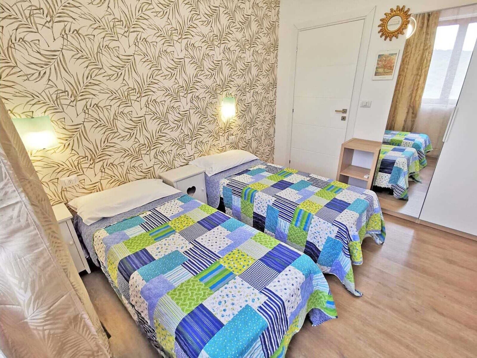Habitació amb dos llits, cadascun amb un estrambó de pegats, paper pintat amb dibuixos, un mirall i una petita estanteria de fusta.