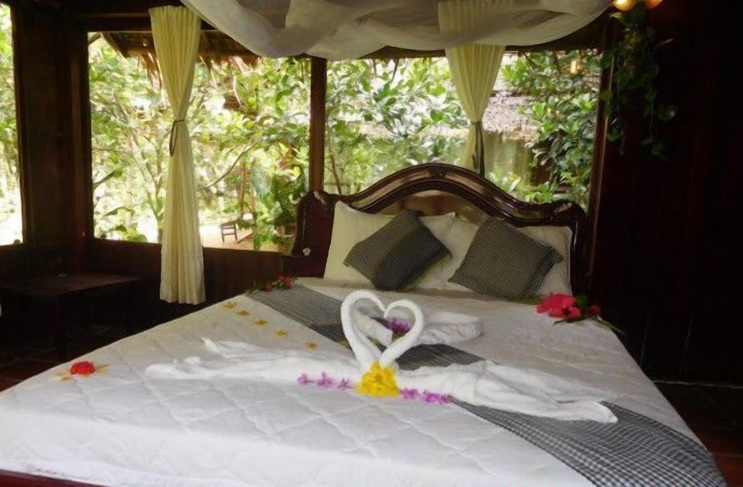 Una cama con una toalla dispuesta en forma de corazón, un dosel y vistas al verdor exterior.