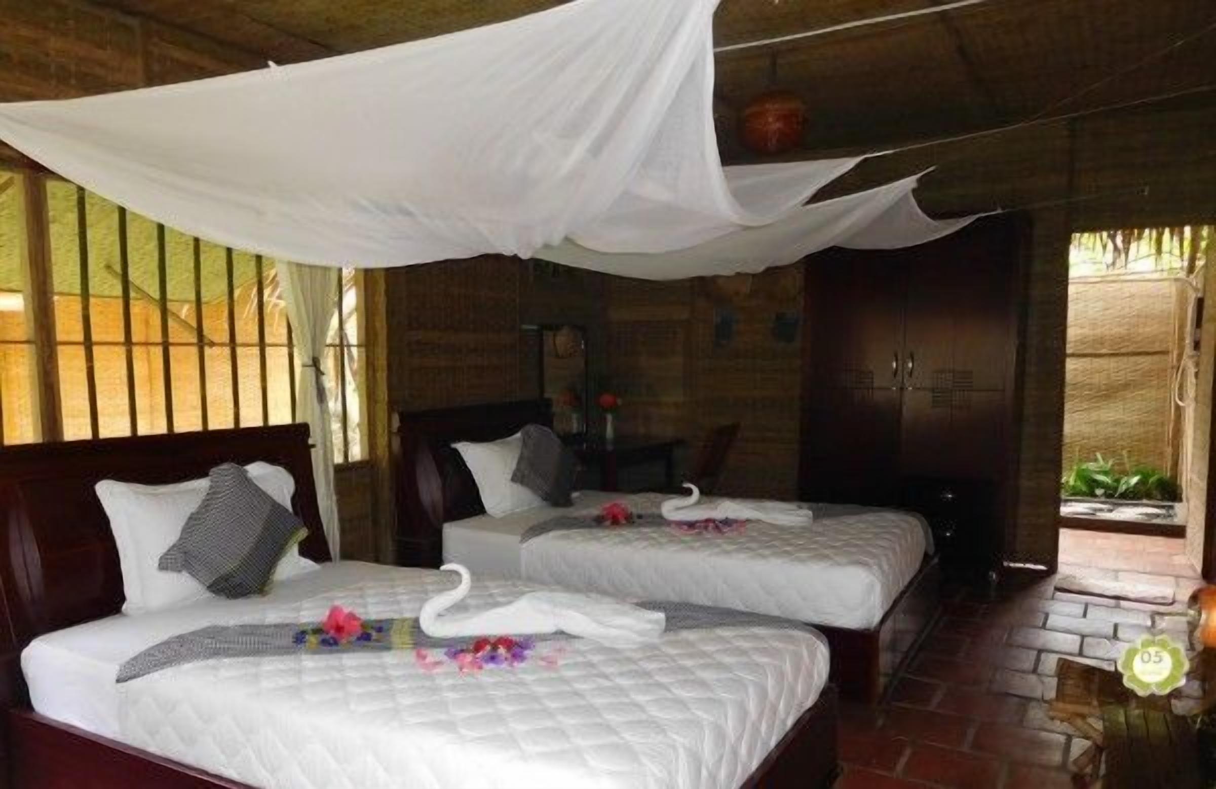 Ein Zimmer mit zwei Betten, jeweils mit weißer Bettdecke und Kissen, und einem Baldachin darüber. Es steht ein kleiner Tisch mit einer Lampe und einer Topfpflanze darin.