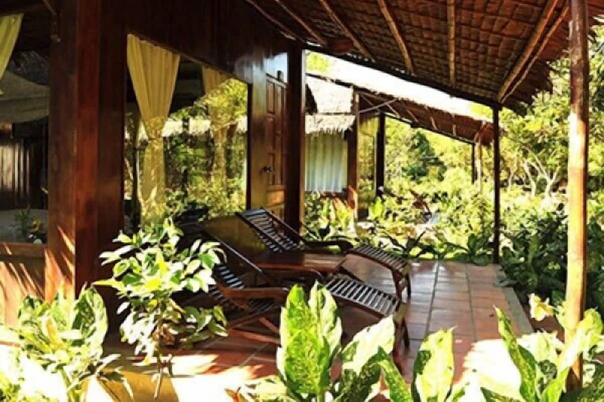 An outdoor covered seating area with wooden furniture, surrounded by lush greenery and a thatched roof structure.