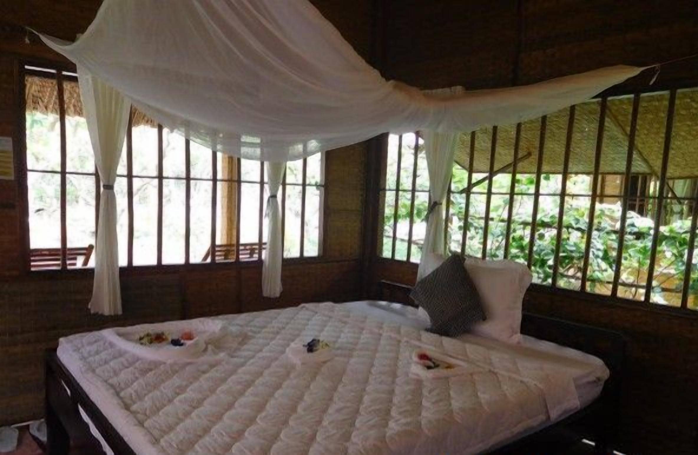 Een kamer met een groot bed, witte gordijnen en uitzicht op groen.