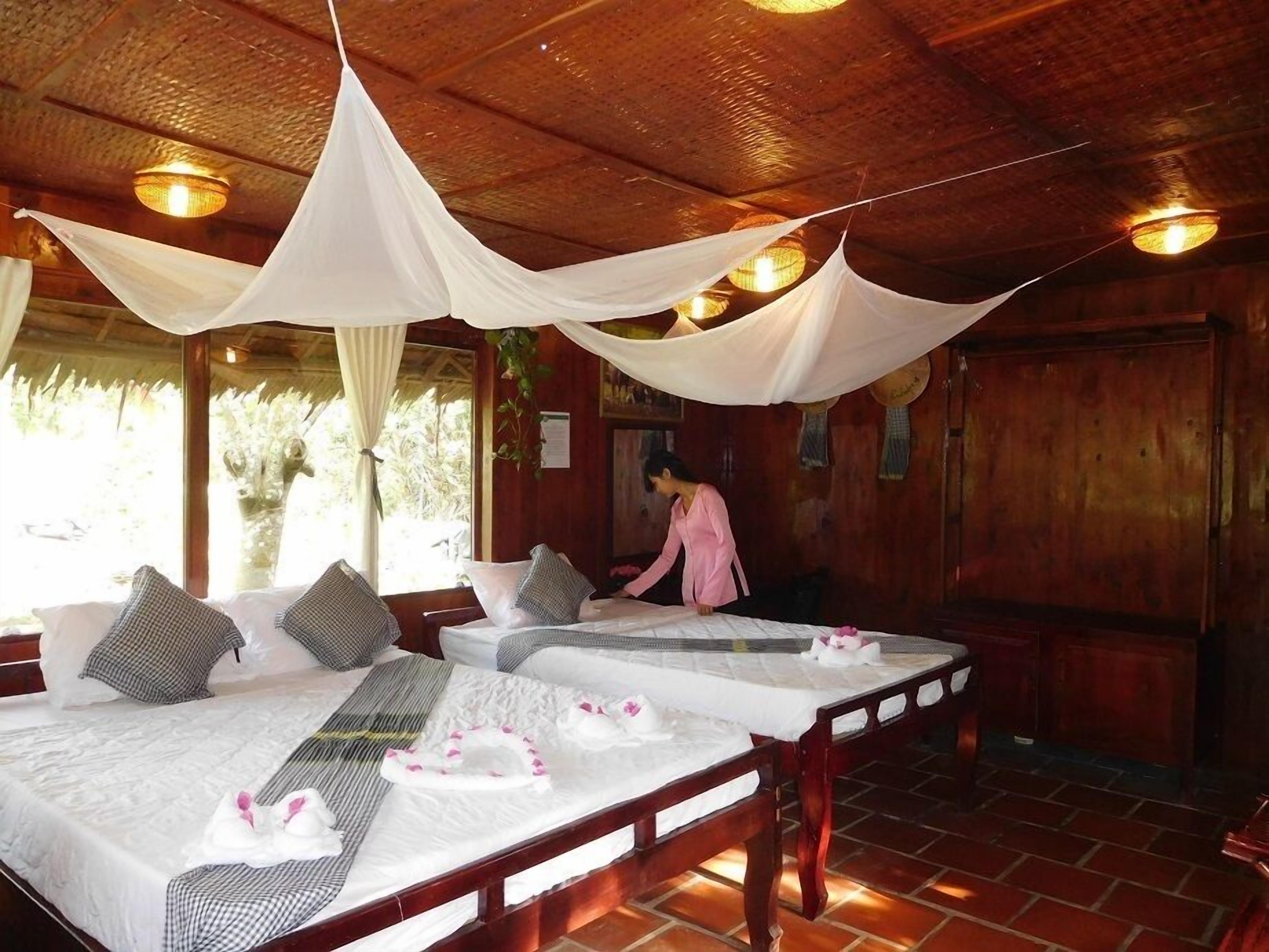 A room with two beds, each with white linens and pillows, a wooden floor, and a person in pink attire.