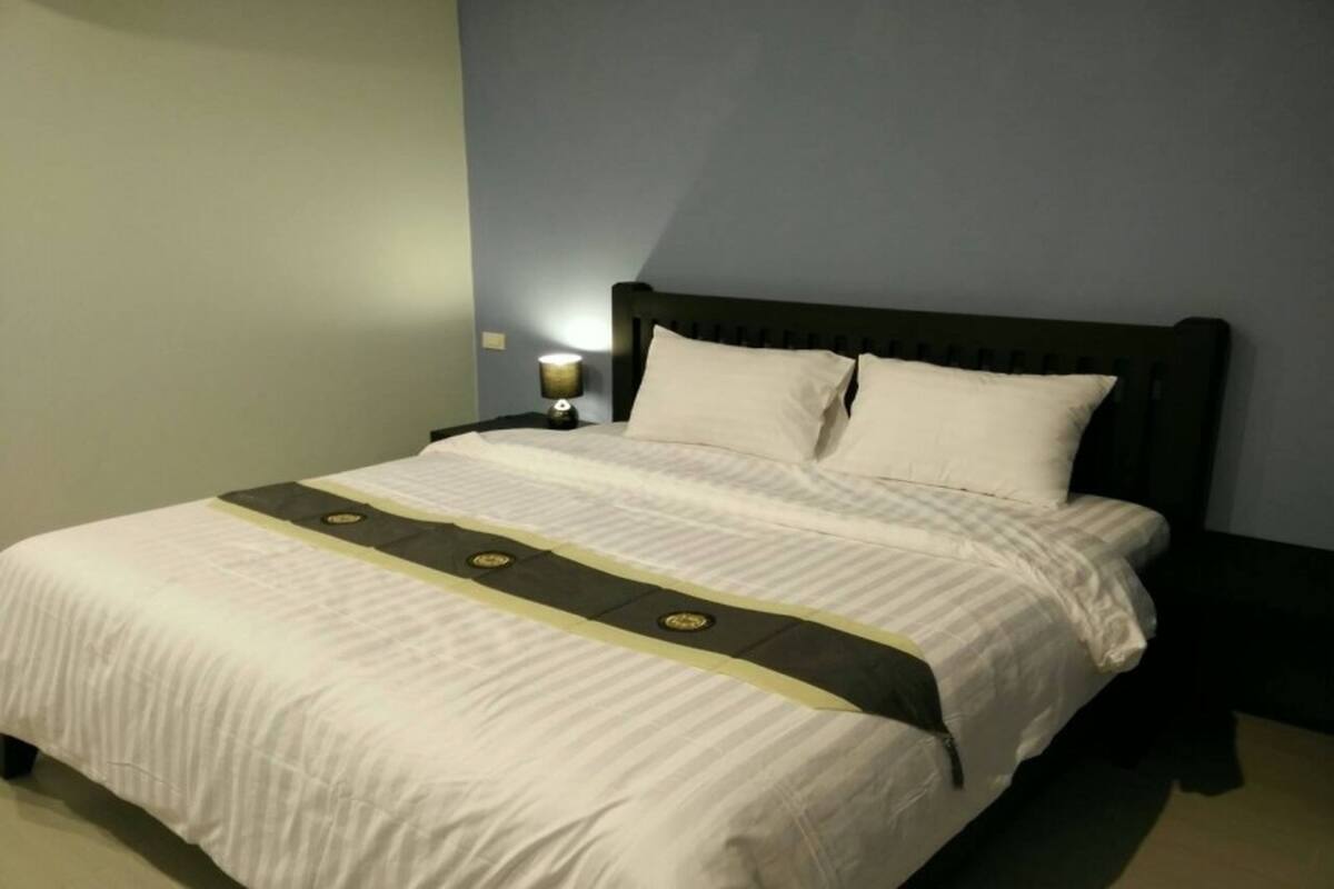 Una cama bien tendida con una colcha blanca y dos almohadas, una mesita de noche con una lámpara y un cabecero oscuro.
