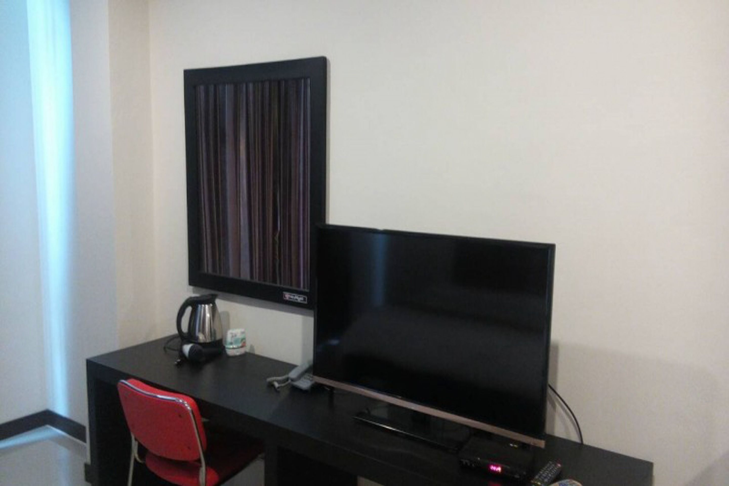 A room with a black desk, a red chair, a flat-screen TV, a coffee maker, and a wall-mounted panel.