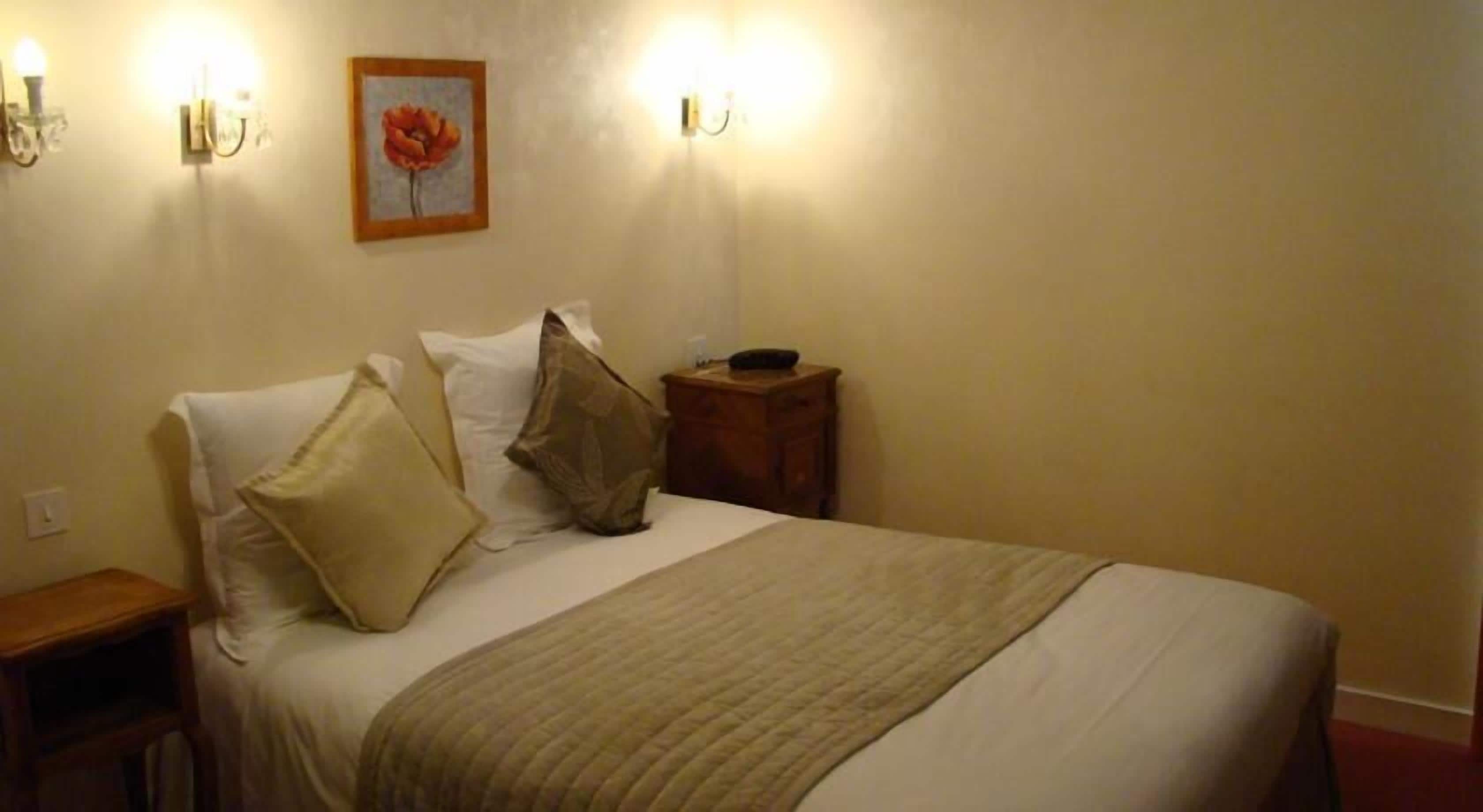 A neatly made bed with a beige blanket, two pillows, and a wooden nightstand with a small table beside it.