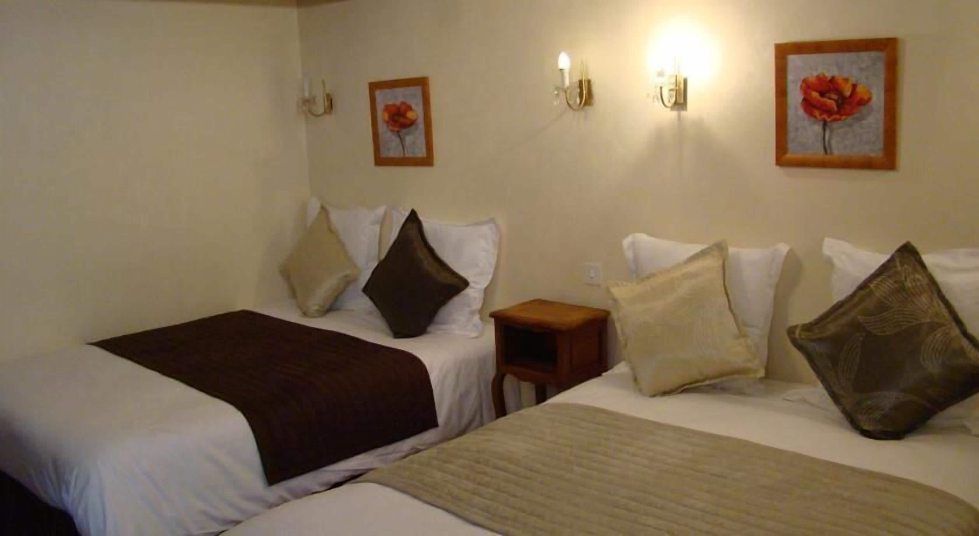 Habitación de hotel con dos camas, cada una con ropa blanca y varias almohadas. Hay apliques de luz en la pared y dos cuadros enmarcados con motivos florales.