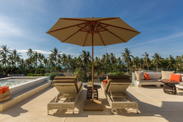 Een zonnig terras buiten met lounge fauteuils, een tafel en een parasol, met uitzicht op een zwembad en palmbomen.