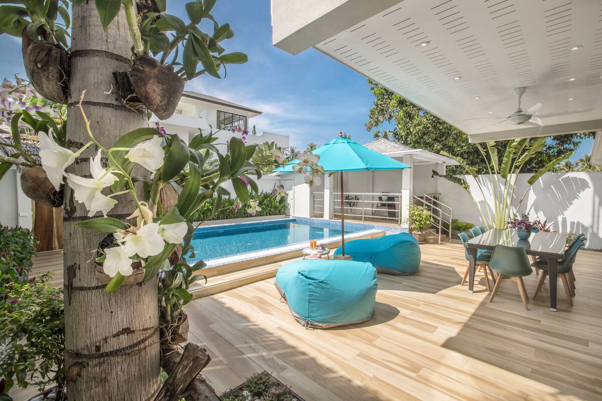 Un espace aménagé au bord de la piscine, avec une terrasse en bois, un coin salon couvert et une table à manger avec des chaises.