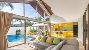 A modern living room with a large grey sofa, a glass door leading to a pool, and a bright yellow kitchen in the background. -  Ban Tai Estate Villas (Koh Samui)