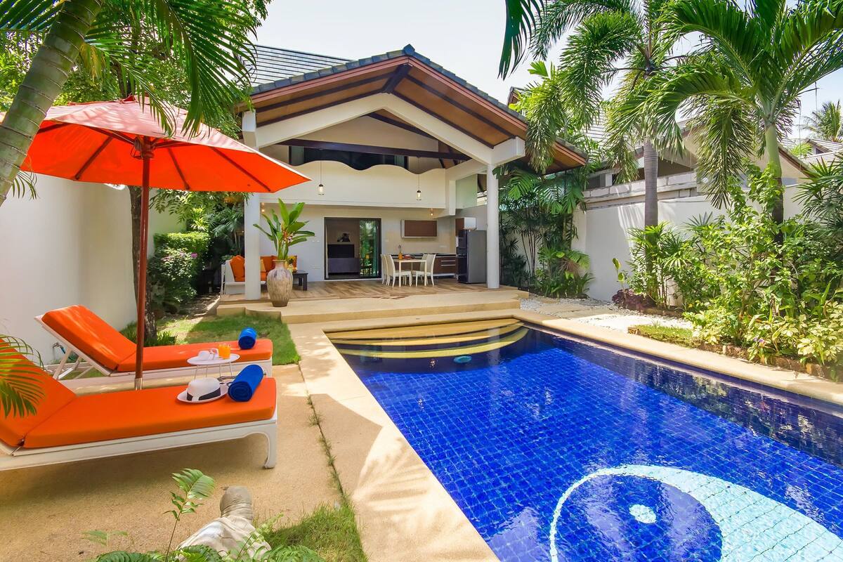 Une piscine entourée d’une végétation luxuriante, avec des chaises longues et un parasol.