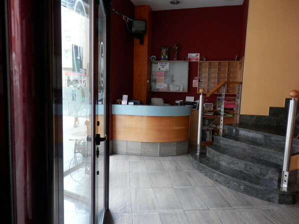 Zona de recepción con mostrador, tablón de anuncios y escalera que sube a un piso superior.