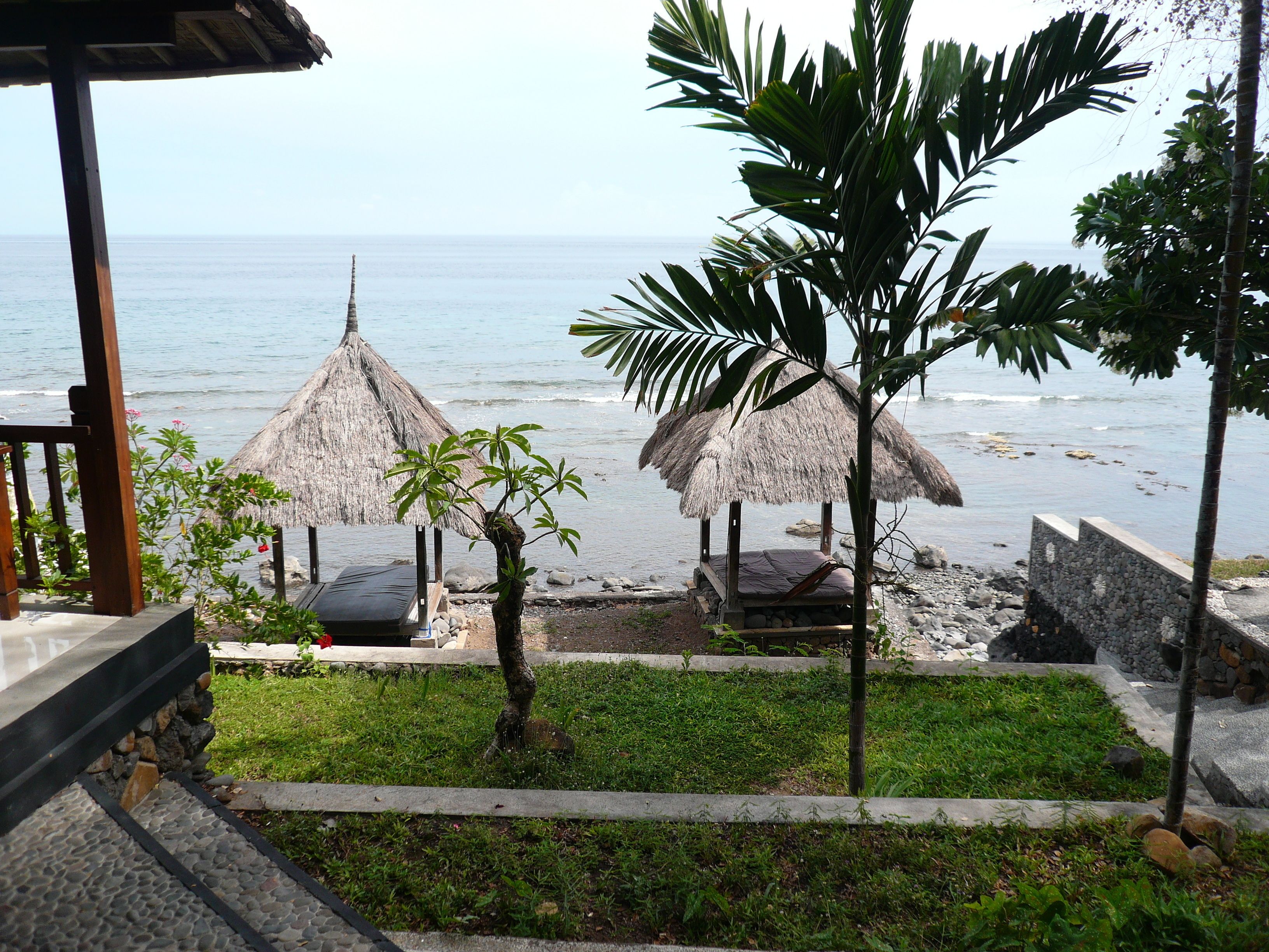 A beachside view with thatched-roof structures, a palm tree, and a stone pathway leading to a seating area.