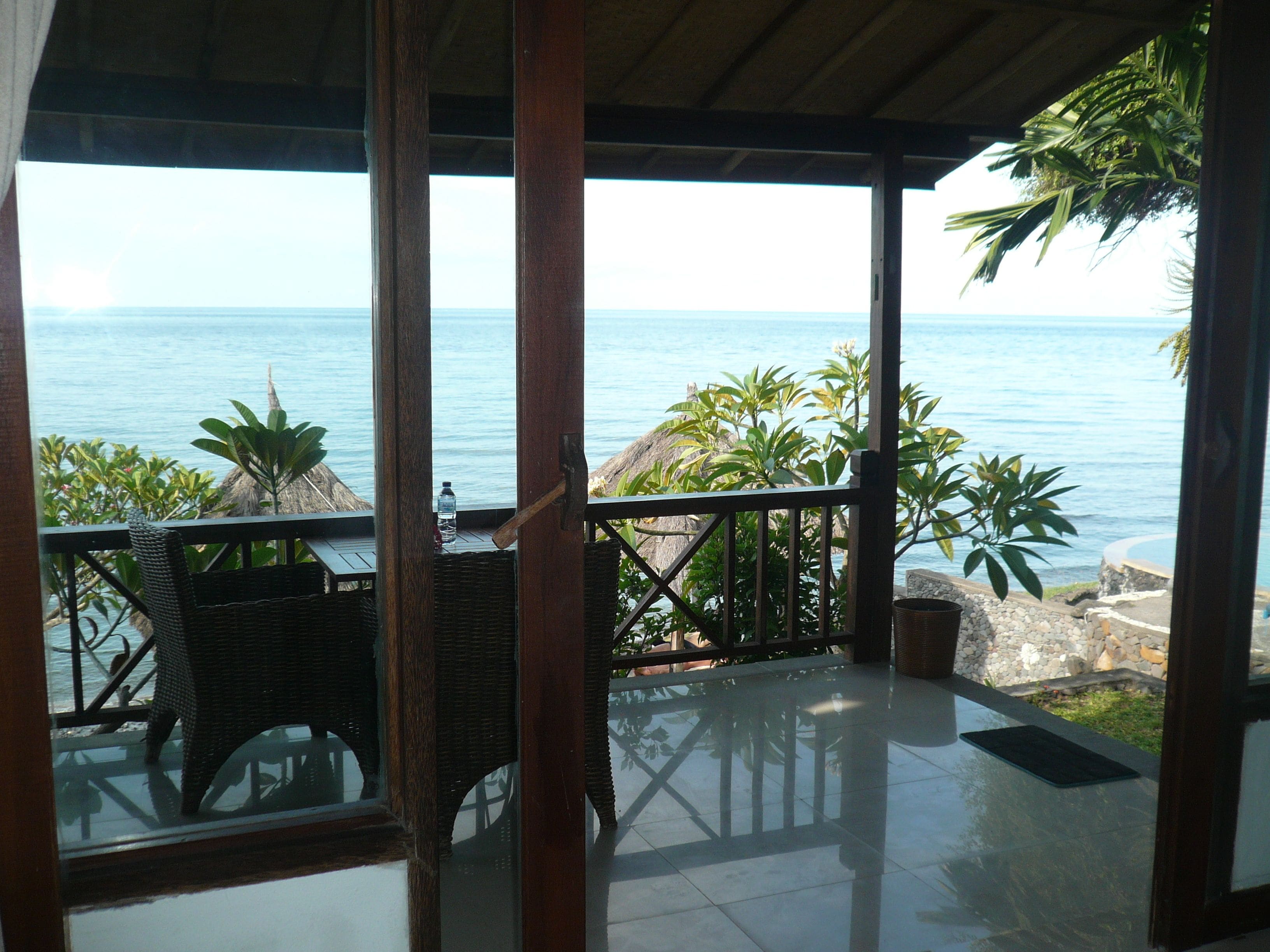 A covered outdoor area with a view of the sea, featuring wicker furniture and a thatched-roof structure.