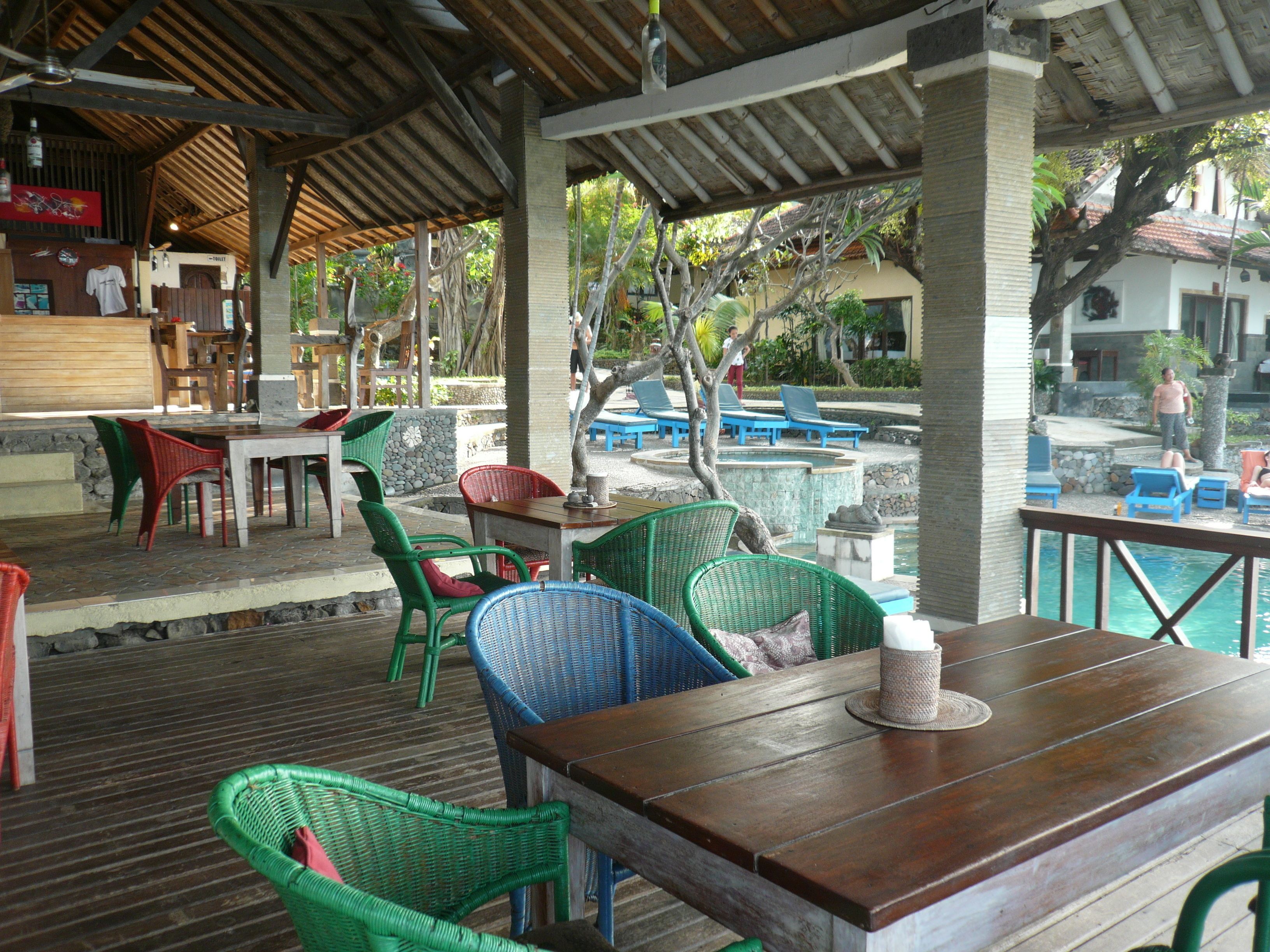 An outdoor seating area with wooden tables and wicker chairs, overlooking a pool and a tropical garden.