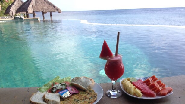 Una mesa de comedor con platos de comida y una bebida junto a una piscina, con vista al océano.