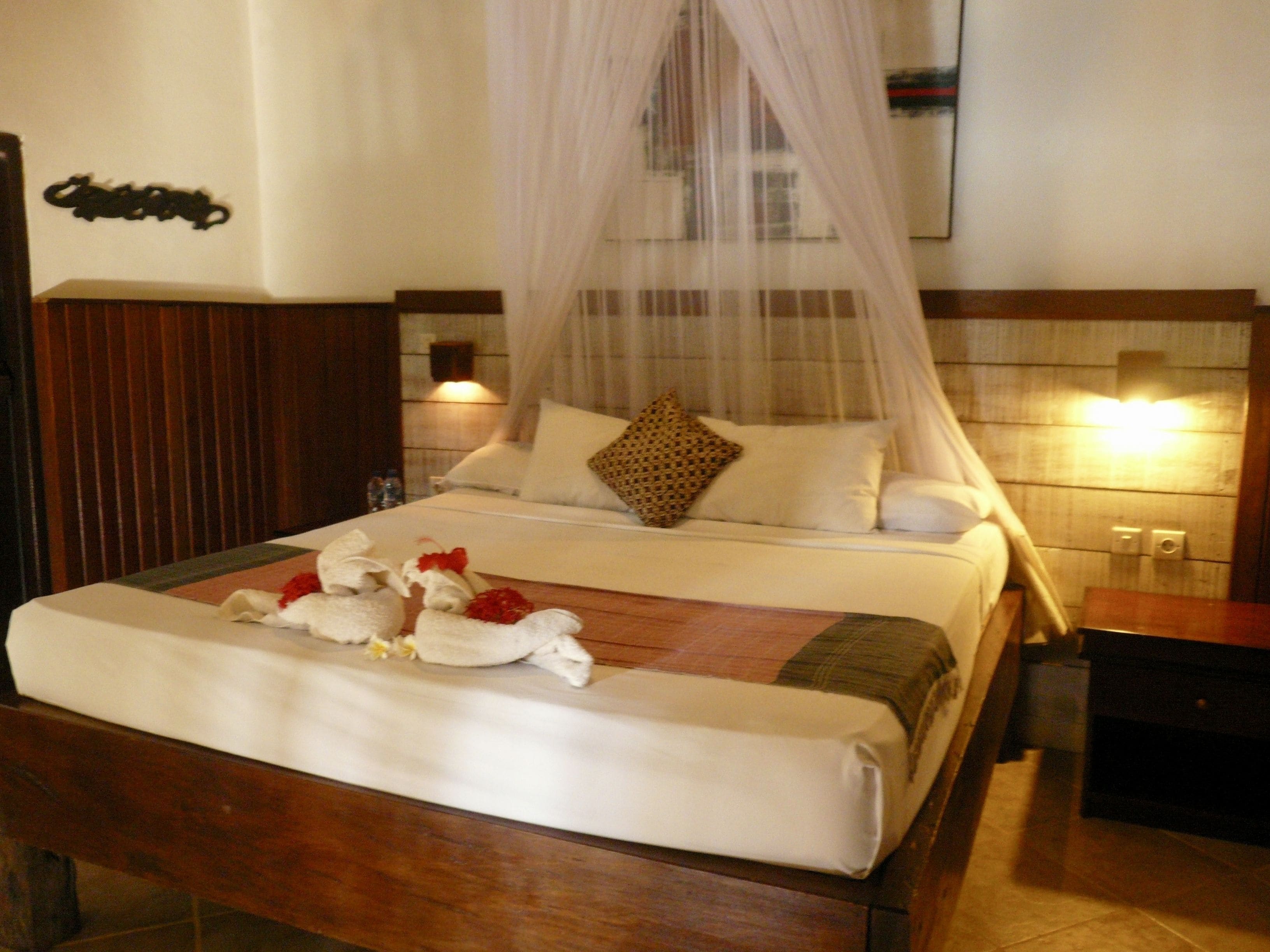 Una cama bien hecha con una colcha blanca, un cojín decorativo y un ramo de flores.