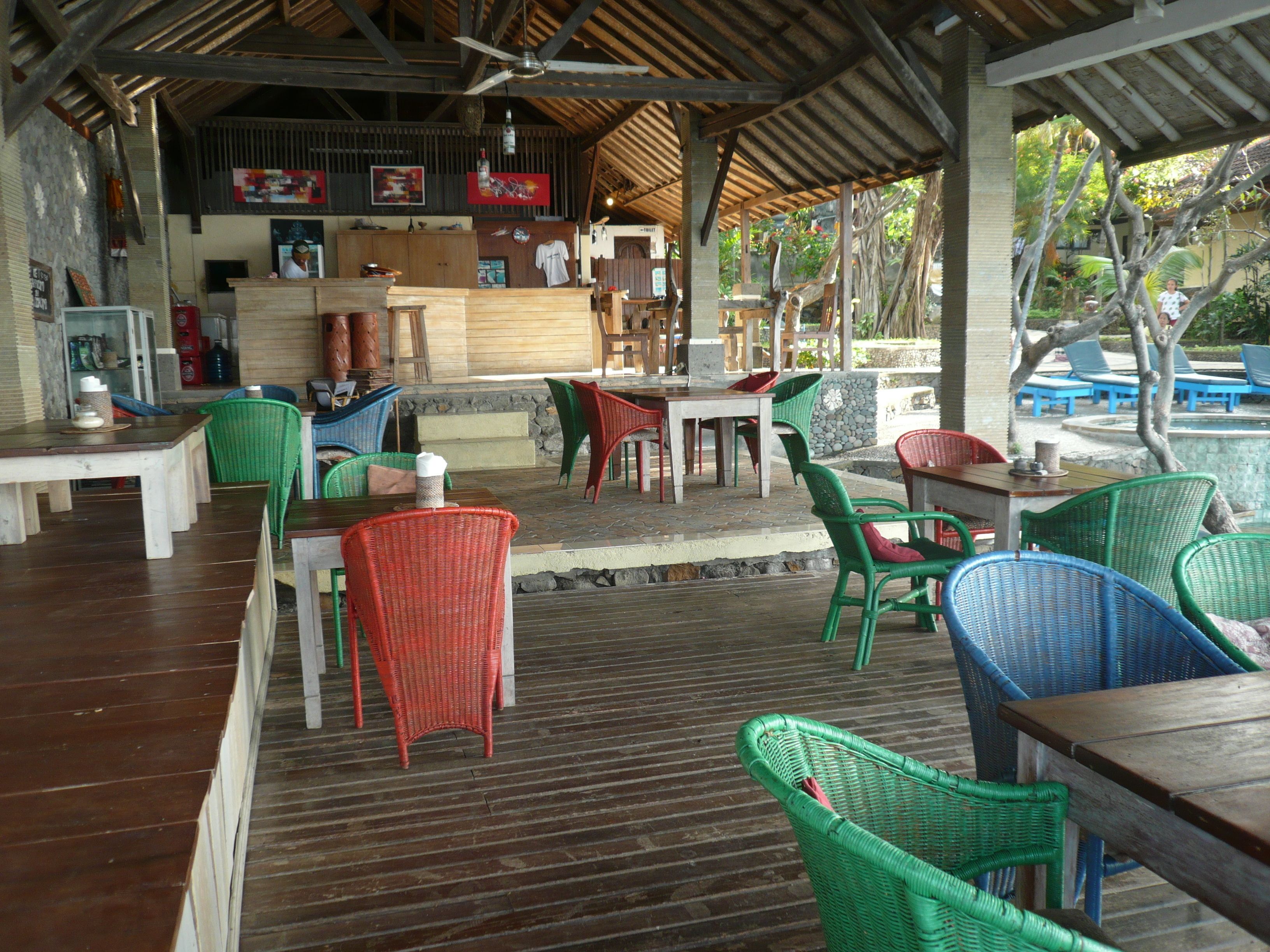 An outdoor restaurant with wooden tables and wicker chairs, a bar area, and a stone wall.