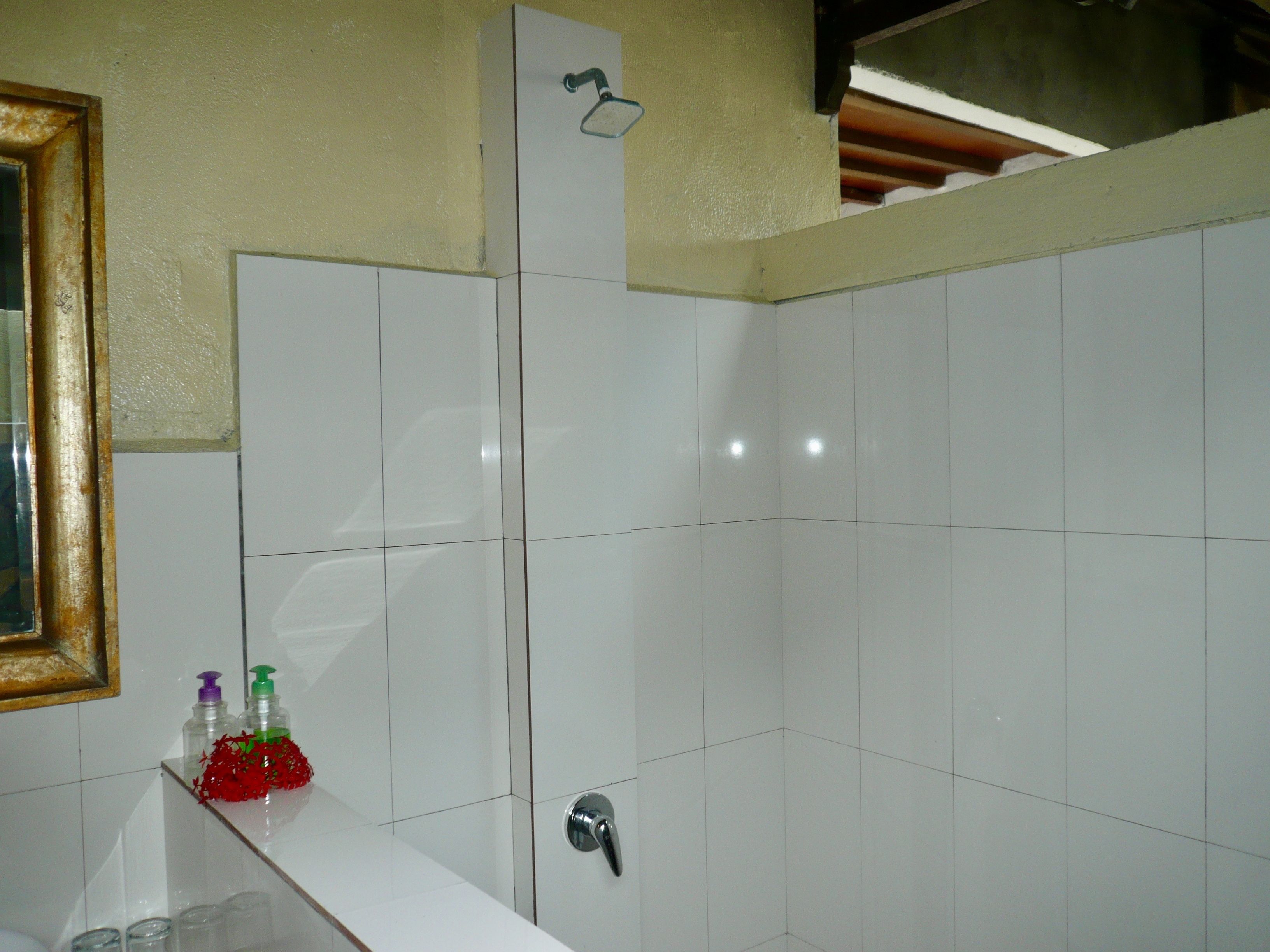 A bathroom with white tiles, a shower, a mirror, and a shelf with toiletries.