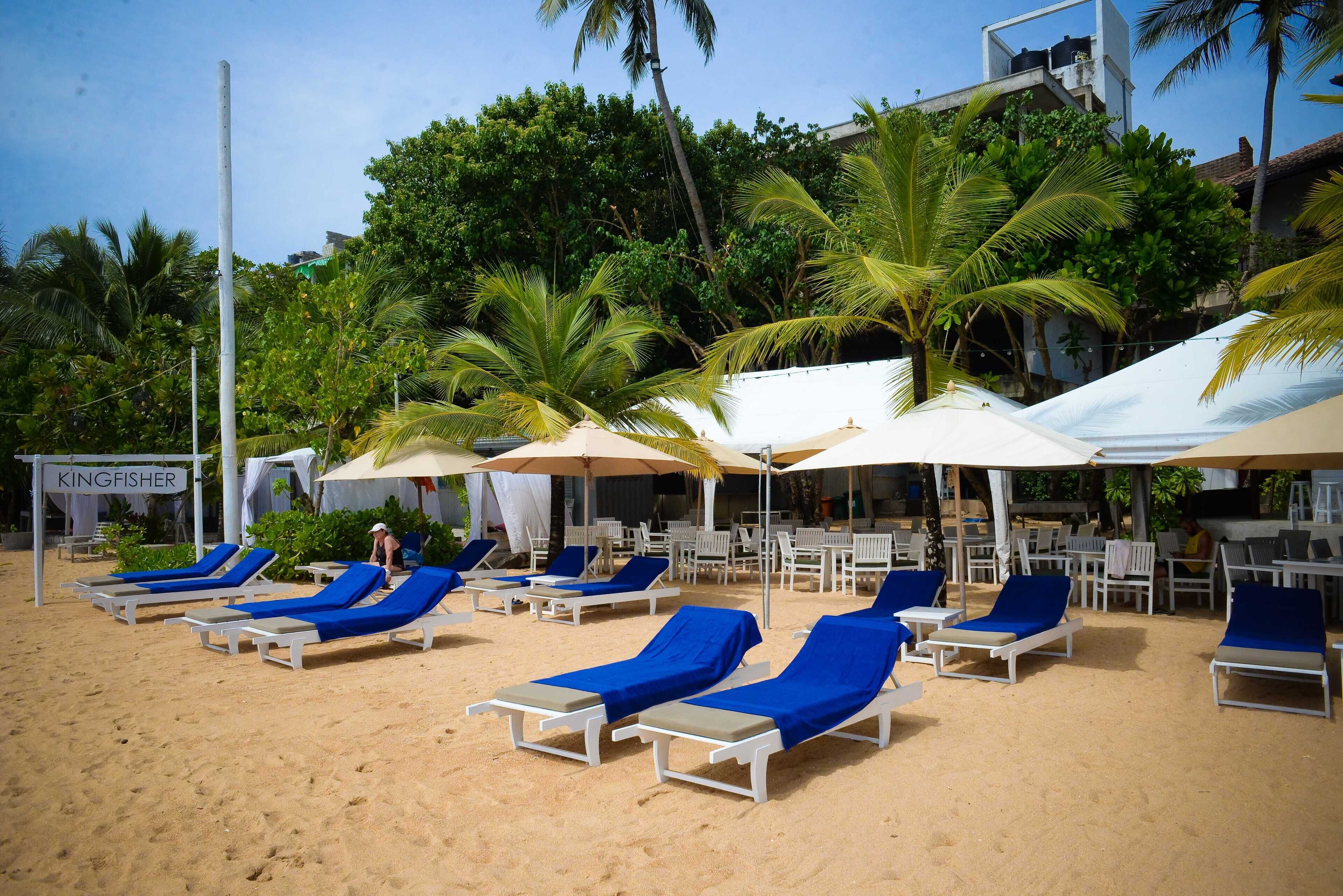 A beachside lounge area with white sunbeds and umbrellas, surrounded by palm trees and a sign reading "KINGFISHER".