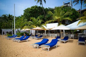 Un espace de détente aménagé sur la plage, avec des chaises longues et des parasols blancs, entouré de palmiers et agrémenté d’une enseigne indiquant « KINGFISHER ».